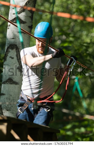 Rope adventure - smiling man\
walking on the rope bridge attached to the tree with an insurance\
hook