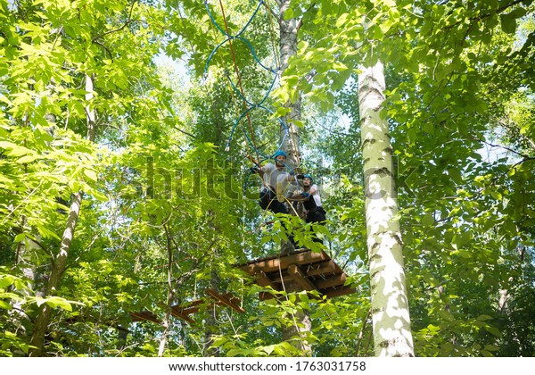 Rope adventure - woman and man having a rope\
entertainment in the green forest - standing on high and walking on\
the rope bridge