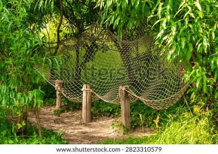 Rope hammock net stretched between wooden posts in lush green park. Outdoor relaxation area surrounded by trees, peaceful recreation, leisure and nature escape