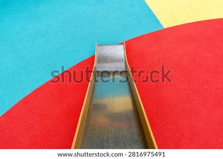 Rope net climbing structure with orange poles over colorful geometric rubber flooring, swings in background, outdoor playground, no people
