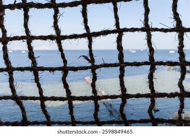 Rope netting frames serene view of coastal resort. Calm blue sea stretches to horizon under clear sky. Natural textures and nautical elements enhance sense of relaxation