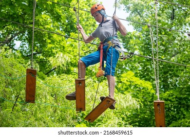 Rope park. A boy teenager in a helmet walks on suspended rope ladders. Carabiners and safety straps. Safety. Summer activity. Sport. Children's playground in nature in the forest. - Powered by Shutterstock