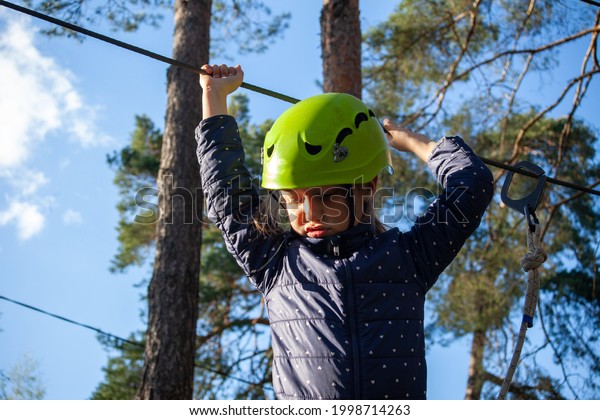 a rope park in a green forest\
recreation park a little girl a school-age child in a helmet and\
with a safety harness goes the distance of a rope\
camp