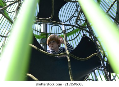 Rope Slide For Small Children. A Little Boy Climbs Up The Ropes. Child Boy Climbed On Top Of The Rope Web On Playground. Kids Play And Climb Outdoors On Sunny Summer Day.