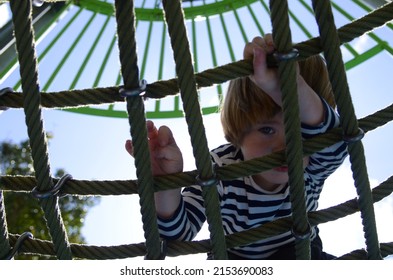 Rope Slide For Small Children. A Little Boy Climbs Up The Ropes. Child Boy Climbed On Top Of The Rope Web On Playground. Kids Play And Climb Outdoors On Sunny Summer Day.