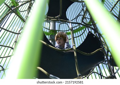 Rope Slide For Small Children. A Little Boy Climbs Up The Ropes. Child Boy Climbed On Top Of The Rope Web On Playground. Kids Play And Climb Outdoors On Sunny Summer Day.