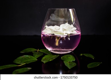 A Rose Flavoured, Pink, Gin And Tonic, Garnished With A Whole Iceberg Rose, On A Black Countertop Strewn With Rose Leaves