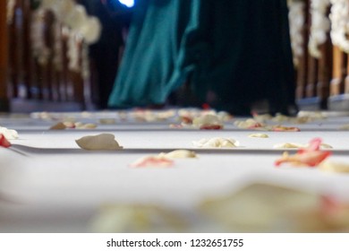 Rose Petals Left On The Wedding Isle After A Wedding Ceremony Has Ended. Bottom Of A Woman's Long Green Gown Is Shown Walking Out The Door With The Wedding Train Of Guests Existing The Church.       