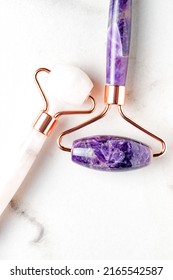 Rose Quartz And Amethyst Face Rollers On Marble Background, Top View. Face Massage Tool, Beauty Treatment And Facial Skin Care Concept.