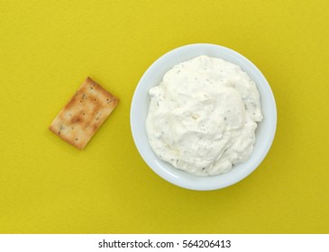 A Rosemary And Olive Oil Seasoned Cracker Next To A Bowl Of French Onion Dip On A Bright Yellow Tablecloth.