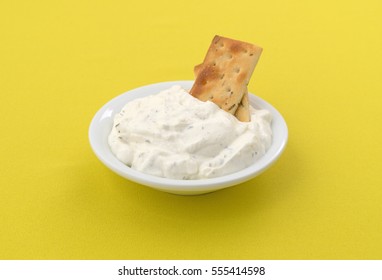 Rosemary And Olive Oil Seasoned Crackers Inserted Into French Onion Dip In A Small Bowl On A Bright Yellow Tablecloth.