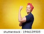 Rosie Riveter concept: proud senior woman with red headscarf, flexing her muscles, copy space, yellow background