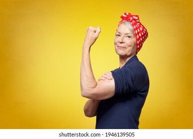 Rosie Riveter concept: proud senior woman with red headscarf, flexing her muscles, copy space, yellow background - Shutterstock ID 1946139505