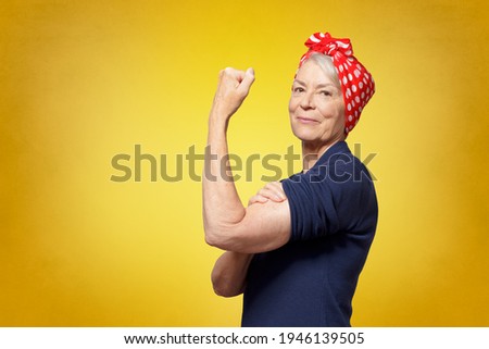 Rosie Riveter concept: proud senior woman with red headscarf, flexing her muscles, copy space, yellow background