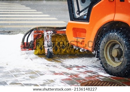 Rotating industrial brush of an orange tractor on a city street during a snowfall. Snow removal, brush machine in winter for clearing the sidewalk. Selective focus.