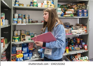 ROTHERHAM, ENGLAND, UK - FEBRUARY 14, 2019: A Young Female Volunteer Working At A Trussell Trust Local Church Food Bank Stands In A Foodbank Warehouse With A Clipboard And Parcel Packing Sheet