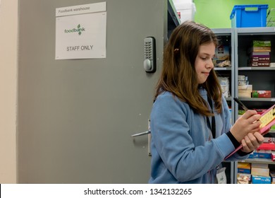 ROTHERHAM, ENGLAND, UK - FEBRUARY 14, 2019: A Young Female Volunteer Working At A Trussell Trust Local Church Food Bank Stands In A Foodbank Entrance With A Clipboard And Parcel Packing Sheet