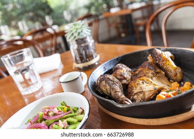 Rotisserie Chicken With Chopped Tomatoes In Iron Pan, With Nopales Salad In White Enamel Bowl, Glass Of Water, Cactus,mug In Restaurant. Dutch Angle