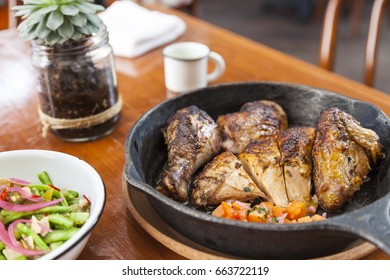 Rotisserie Chicken With Tomatoes In Iron Pan And Nopales Salad In White Enamel Bowl, Mug, Cactus And Chair On The Background.Horizontal
