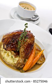 Rotisserie Roasted Chicken Leg Served Over Mashed Potatoes With Baby Carrots And A Gravy Sauce.  A Fresh Green Sprig Of Rosemary Tops This Off.  A Warm Bowl Of Soup Is Featured In The Background.