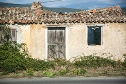Rotten And Deteriorated Old Vintage House With Wooden Door And Windows Somewhere At Corse. Tiled Roof Is Somehow Broken,  Brushwood In Front.  Traces Of Time Are The Witnesses.