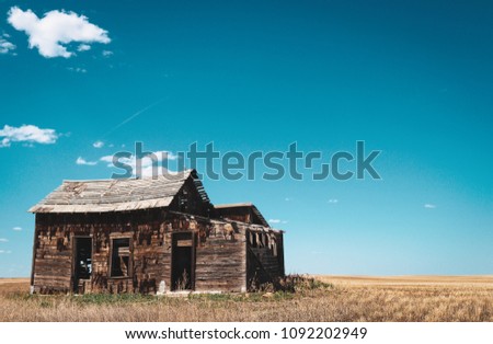 Rotting abandoned farmhouse sits forlorn in a recently plowed field with blue skies and sparse clouds 
