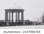 Rotunda on the embankment in winter. Snowing. The transparent dome is covered with snow. Lighthouse in the background in blur. Cloudy day on the city promenade.