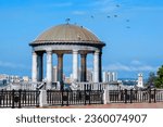 Rotunda on the river embankment against the blue sky. A flock of pigeons on the transparent dome of the gazebo. Lighthouse in the background. Structures on the promenade. Summer sunny day.