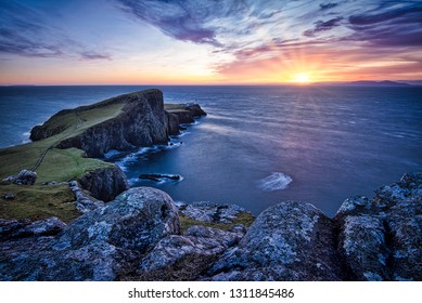 Rough coast, rocks, cliffs, lighthouse and the sea during colorful sunset. Cold scene from the north. Pleasing warm sunset, the sun setting on horizon, rays of light, colorful skies and clouds.