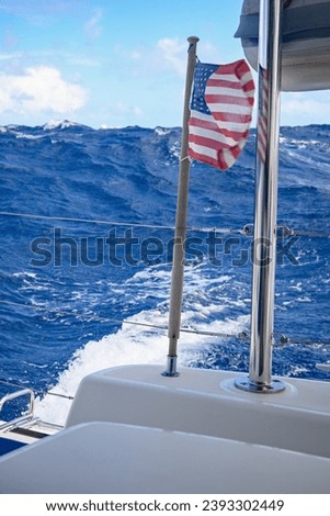 Rough ocean water is swelling behind the stern of a catamaran with the American flag blowing in the wind, underneath the bottom of a dingy.