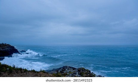 Rough ocean waves break on a rocky coastline under a peaceful, cloudy blue sky