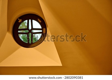 Round attic window with dark frame, golden hardware, and greenery visible outside, set against an ochre angled wall