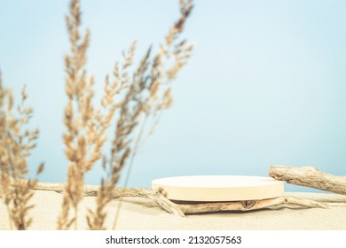 Round beige platform podium, dry tree twigs on white beach sand with dry bent plant in foreground. Minimal creative composition background for cosmetics or products presentation. Front view