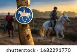 A round blue and white sign marks a bridle path in Spain. Here it is allowed to ride horses. In the background out of focus with bokeh two horsemen and a landscape at sunset.
