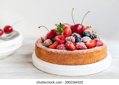 Round cake decorated with fresh summer fruits powdered with sugar, freshly baked homemade pastry topped with strawberries, raspberries, cherries, ready to eat cake served on white wooden table