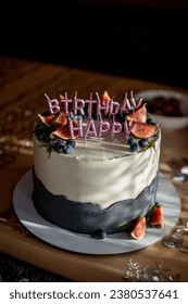 A round cake is on the table. Vertical photo. Candles in the form of letters Happy Birthday. Decorated with berries and fruits, blueberries and dates. White blue color Presents at birthday party.