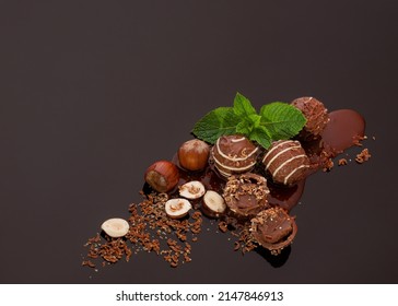 Round chocolate candies with a soft filling scattered in the lower right corner with chocolate chips, nuts and mint leaves on a dark chocolate background with reflection. The upper left corner is empt