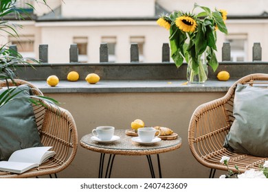 Round coffee table close to two rattan armchairs with cushions at open balcony. Hot tea drink in cups and croissant. Good morning concept. Breakfast in cozy hotel patio with flowers.