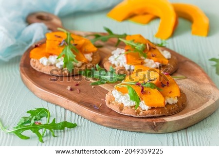 Round crispbread slices with roasted pumpkin, grainy cream cheese and rocket salad on a wooden board on light green background [[stock_photo]] © 
