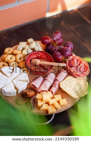 Round cutting board with slicing assorted cheese, sausage rosette, ham, cracker, grapes and figs with honey. Charcuterie and cheese platter on the terrace with sunbeams and palm leaves