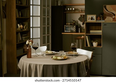 Round dining table set with two plates of assorted cheese and fruit, two glasses of red wine, decorative vase with flowers