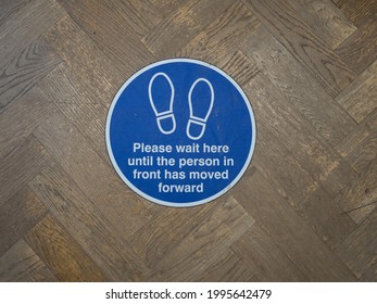 A Round Floor Sign With White Text On Blue Background And Two Footprint Symbols.It Says Please Wait Here Until The Person In Front Has Moved Forward .Coronavirus Pandemic