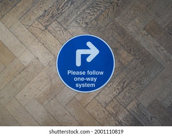 A Round Floor Sign With White Text On Blue Background Has An Arrow Pointing Right.It Says 'please Follow One Way System' .Coronavirus Pandemic