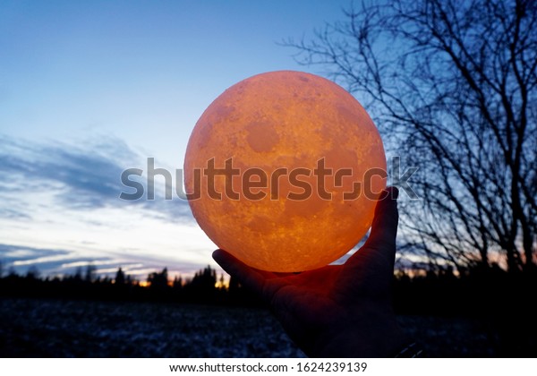 Round full moon in hands\
against evening sky. Lunar model, moon-shaped lamp with moon\
craters         