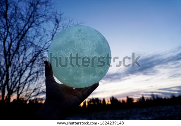 Round full moon in hands
against evening sky. Lunar model, moon-shaped lamp with moon
craters         