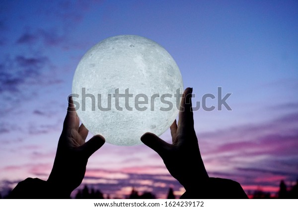Round full moon in hands\
against evening sky. Lunar model, moon-shaped lamp with moon\
craters         
