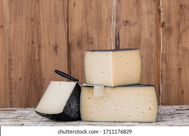 Round Head Of Hard Cheese With Small Cut Slice On Wooden Background Close Up. Farm Homemade Cheese. The Concept Of Nutrition And Healthy Lifestyle. Heads Of Young Goat Cheese With Black Shell.