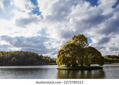 A round island with trees growing on it in the middle of a lake backlit by the sun on a cloudy day in autumn in Tsaritsyno Park, Moscow, Russia - Shutterstock ID 2275789213