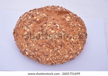 A round loaf of multigrain bread sits on a light surface. The bread is speckled with seeds and grains, giving it a rustic, textured appearance. It looks wholesome and freshly baked.