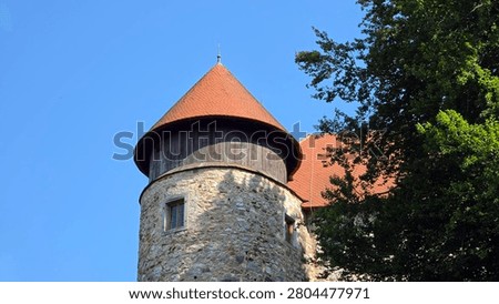 A round medieval guard tower built from textured stone blocks features an overhanging wooden upper section and a red clay tile roof set against a vibrant blue sky